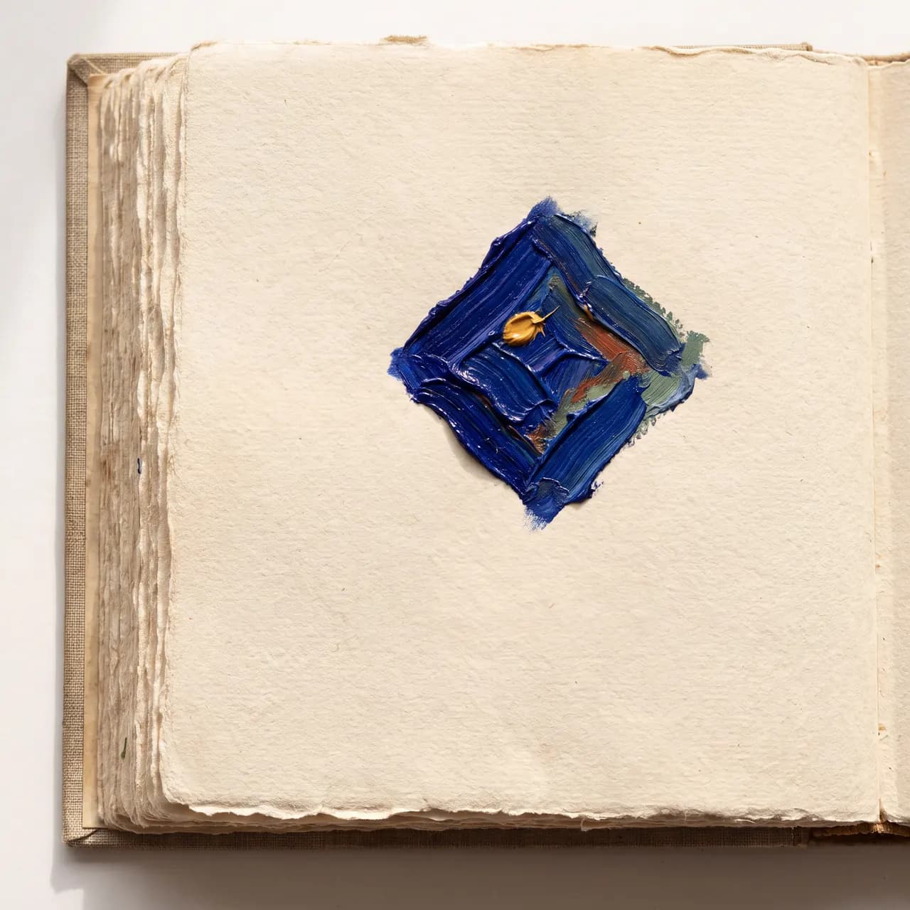 A small, diamond-shaped patch of thick blue and yellow oil paint is centered on the cream-colored, deckled-edge page of an open handmade notebook.